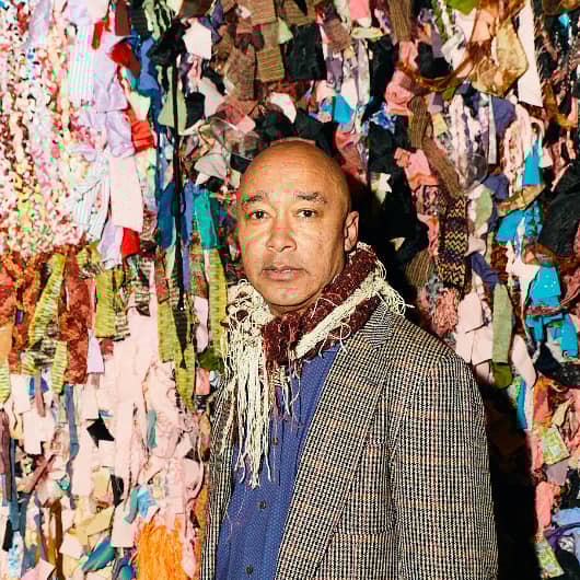 Portrait of the artist Ramekon O'Arwisters in front of one of his fiber art sculptures. He is wearing a tweed jacket, dark blue shirt, and a scarf with lots of fringe. His work is composed of many many strips of colorful fabric loosely woven together.