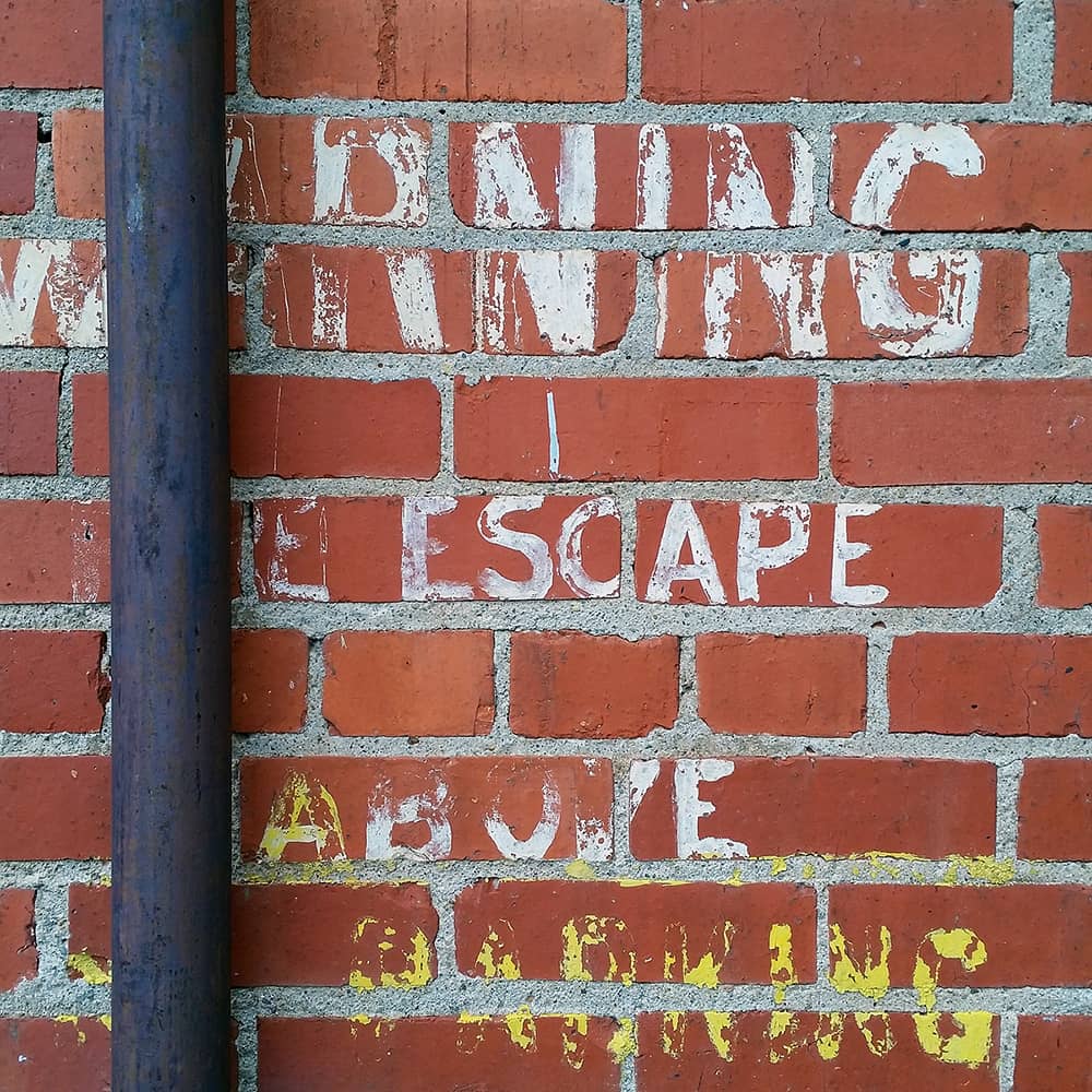 Photo of a brick wall with a dark pipe on the left. The fading paint reads "Warning fire escape above no parking"