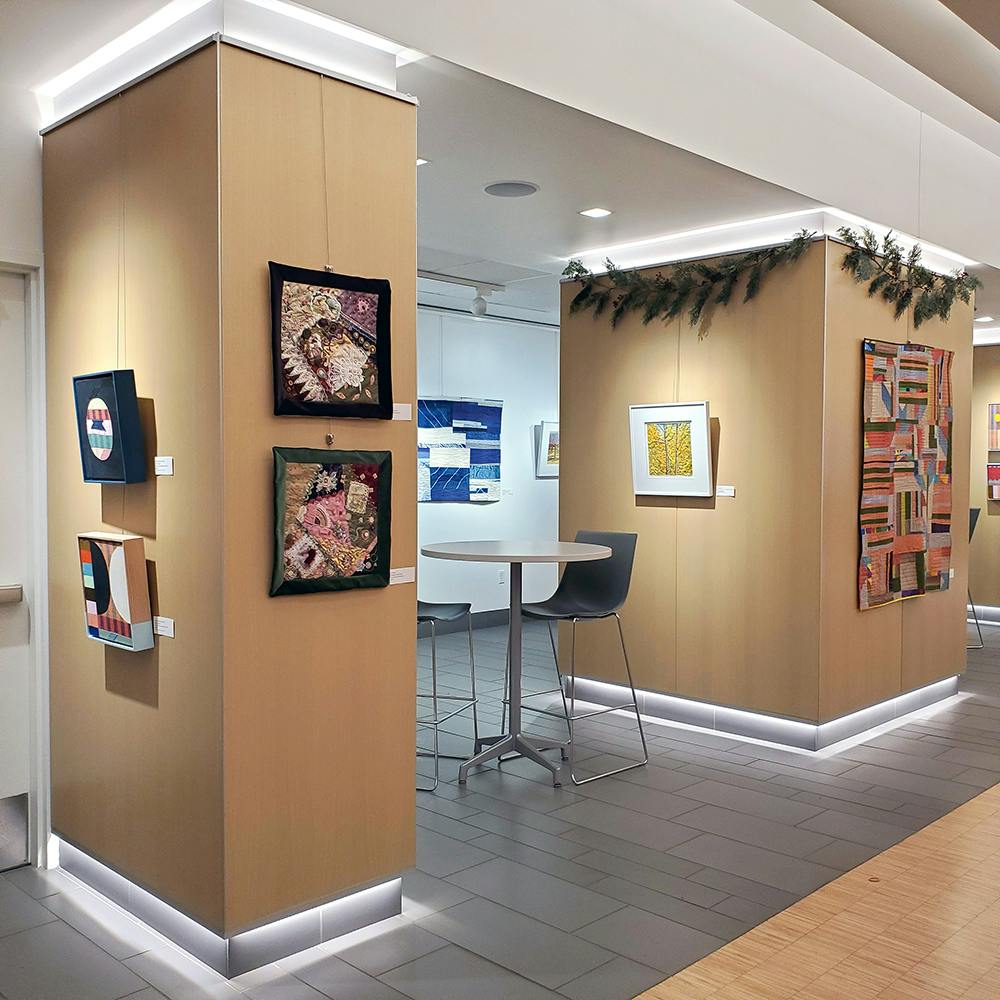 Photo of a gallery space in which quilts and other fiber art pieces are displayed on the walls.