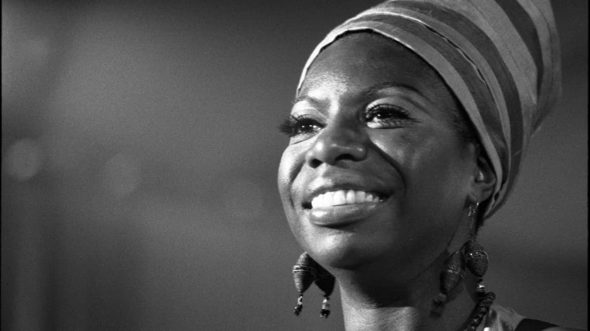 A black and white portrait photo of the musician Nina Simone. We see Ms. Simone from the neck up, smiling, wearing a cloth head wrap and large beaded earrings.