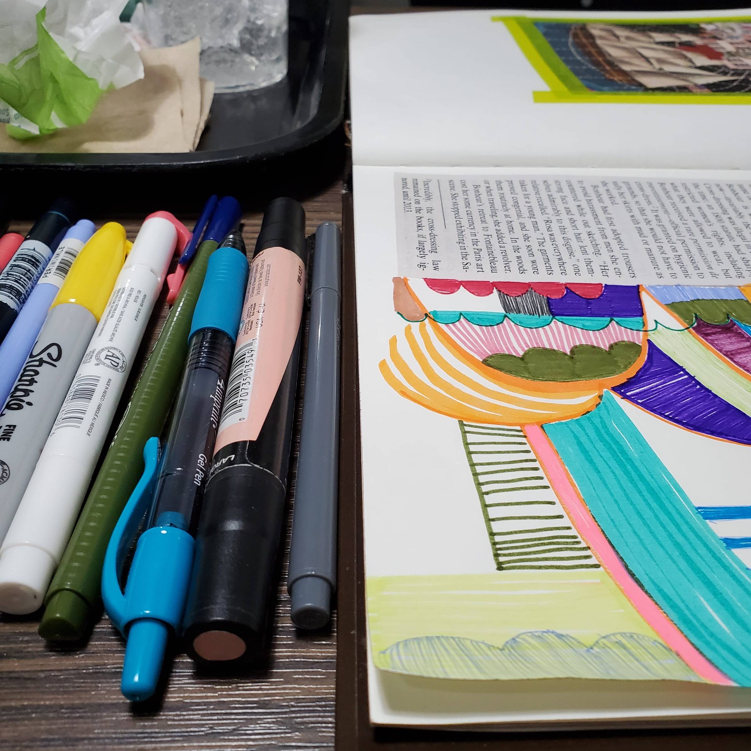 Close up photo of a sketchbook with multicolored doodles in it. To the left of the sketchbook are several colored pens. In the background is a black plastic tray containing taco wrappers.