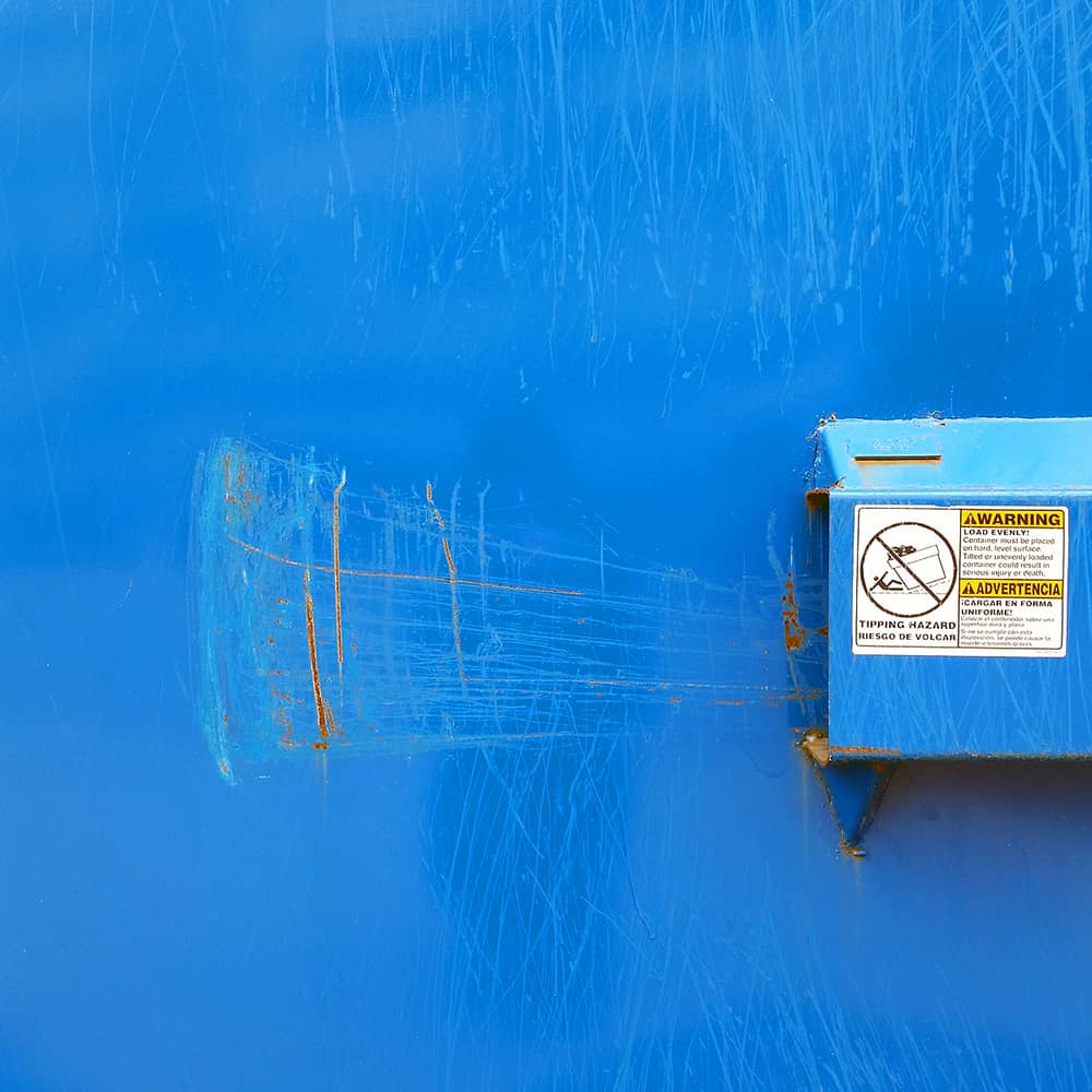 Photo of the side of a bright blue dumpster. The blue paint shows multiple scrapes and rust. To the right is a sticker warning of tipping hazard.