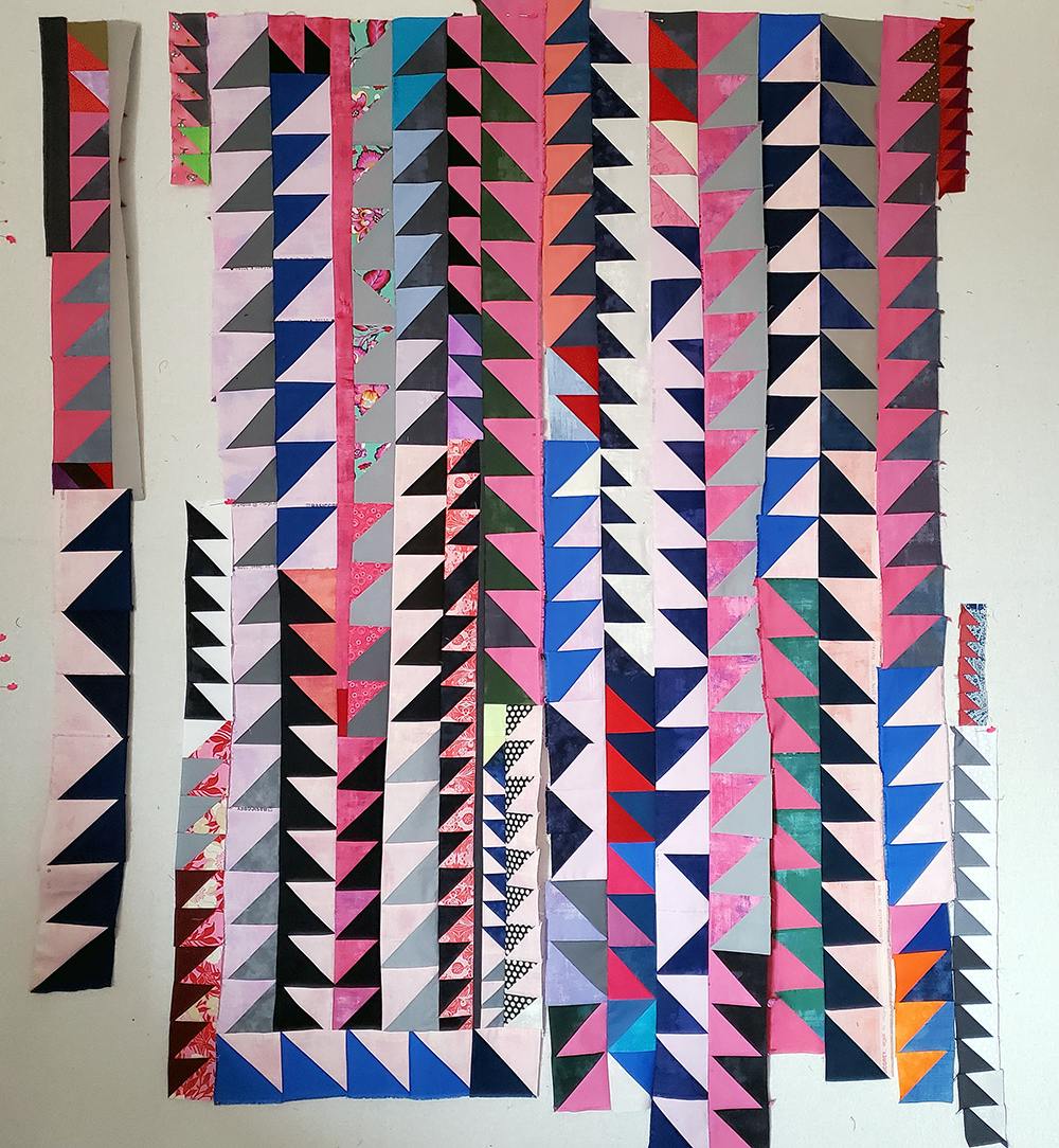 Photo of a patchwork quilt in progress against a white background. The fabric pieces are mostly triangular, in various shades of pink, gray, and blue.