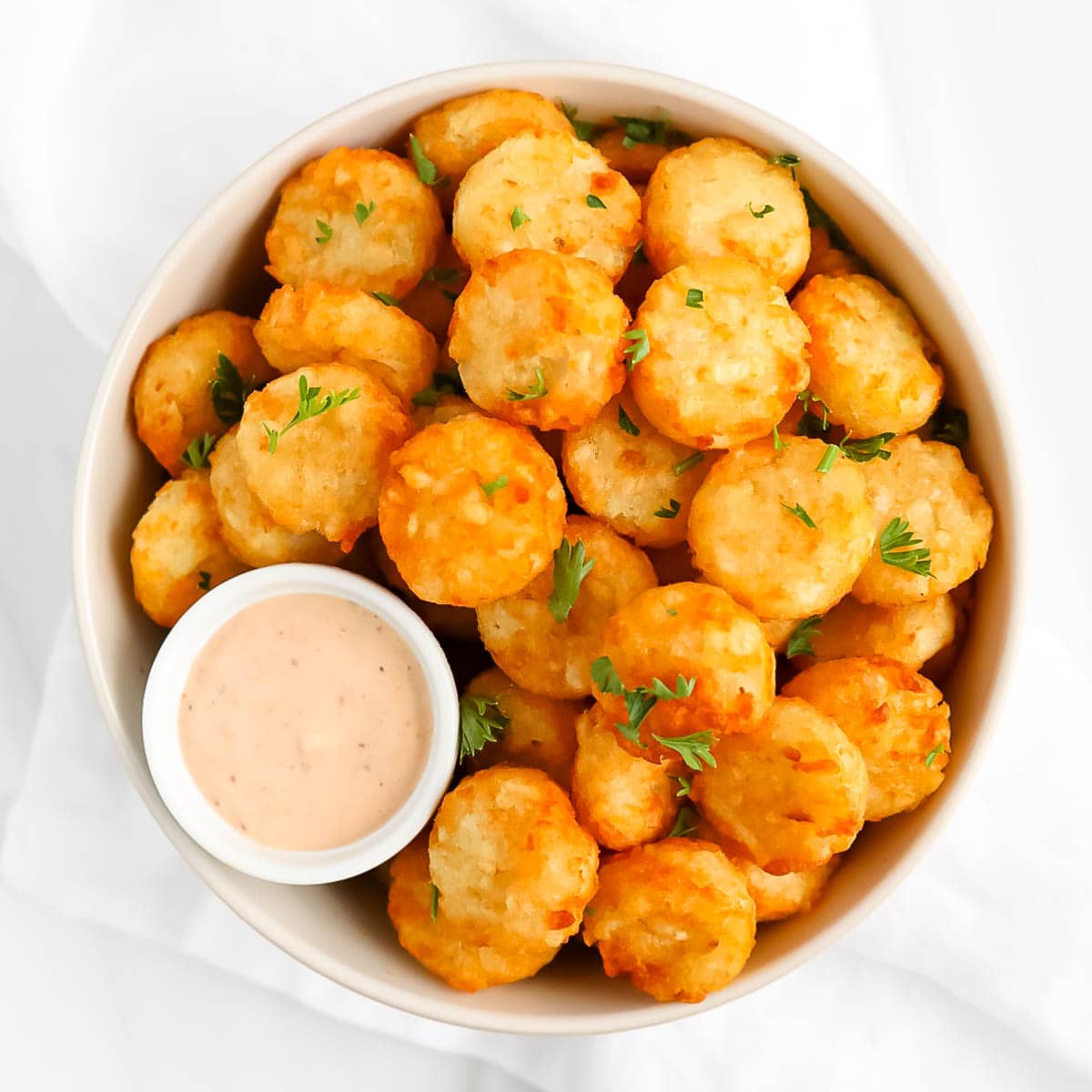 Photo of crispy crowns, which are fried potato nuggets, in a bowl with a smaller bowl of sauce. The golden potato pucks are garnished with chopped parsley. This image was sourced from the cooking blog https://plantedinthekitchen.com/.