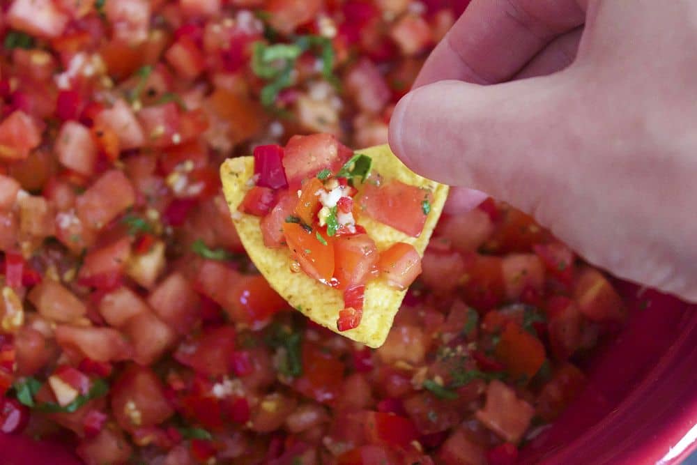 Photo of a caucasian hand holding a triangular tortilla chip. The chip holds a spoonful of tomato-based salsa. In the background there is a large amount of the salsa in a red bowl.