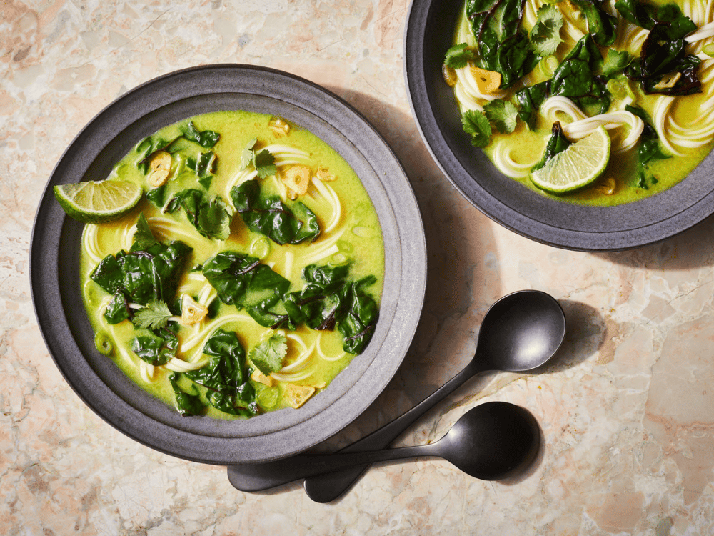 Photo of two bowls of ginger turmeric soup sitting on a marble countertop. The soup is a light green color, and includes noodles, dark green leaves, and slices of green onion and lime. There are two soup spoons to the right of the lower bowl.