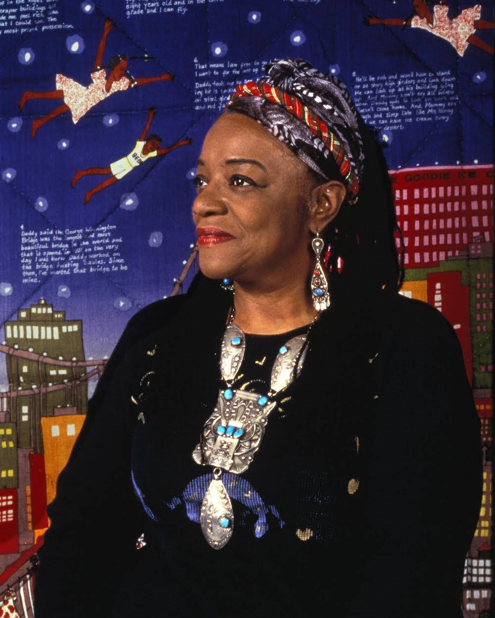 A portrait photograph of artist Faith Ringgold which appeared along with her 2019 retrospective exhibition in London. Ringgold is an African-American woman, dressed here in a black outfit with large silver jewelry and multicolored hair wraps. She is posed, looking to her right, in front of one of her most famous works, Tar Beach.
