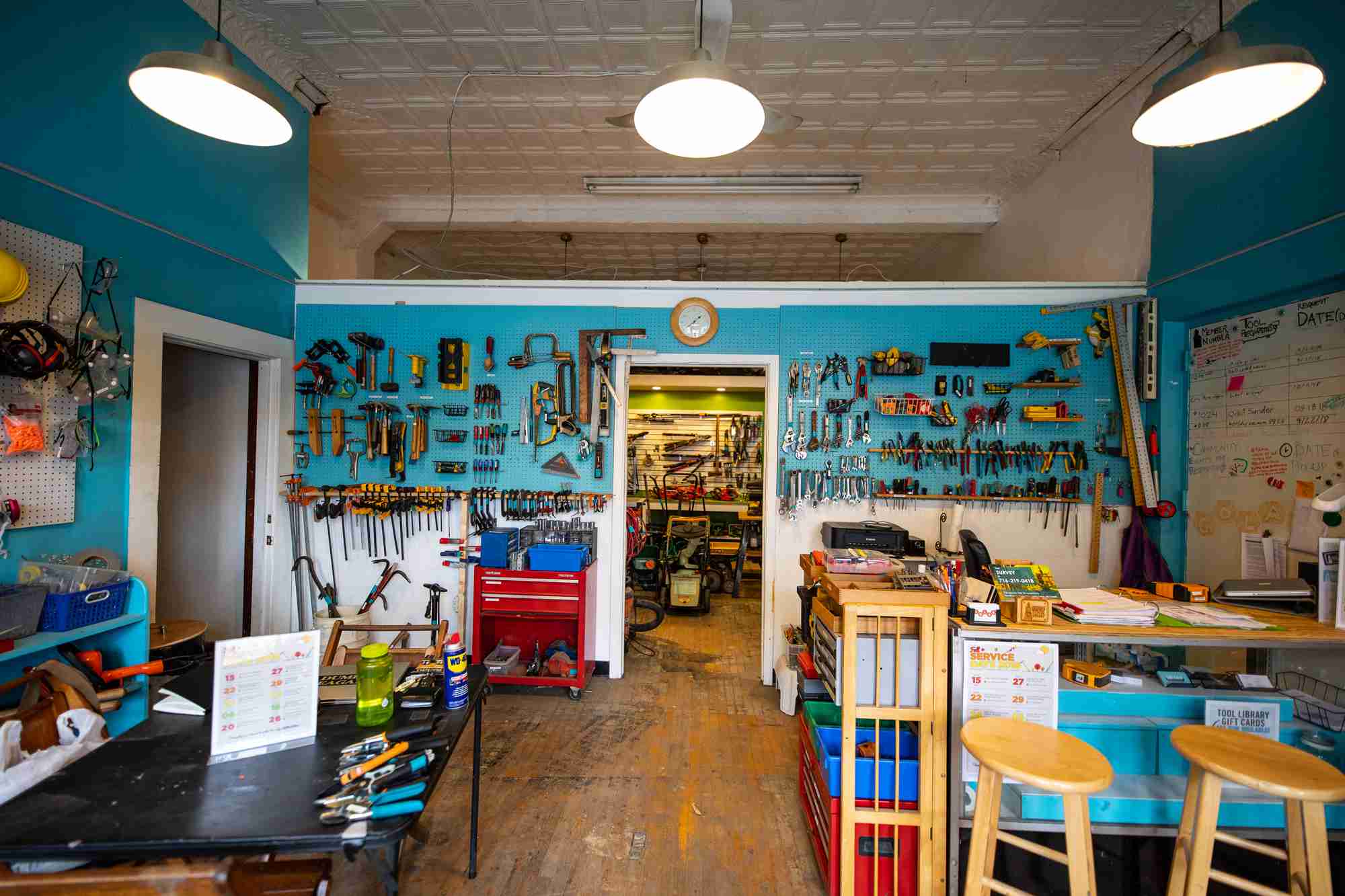 Photo of an orderly interior of a workshop where multitudes of tools are available for people to check out. The room has a wood floor, white tile ceiling, and walls painted cerulean blue. The walls have many tools hanging on them. Several tables and carts hold more tools.