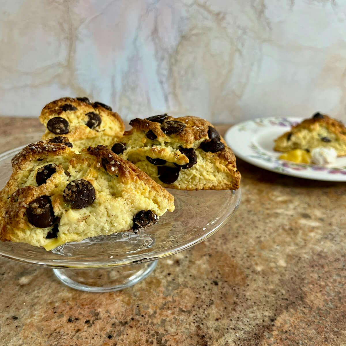 Chocolate chip scones on a cake stand with one on a flowered plate behind.