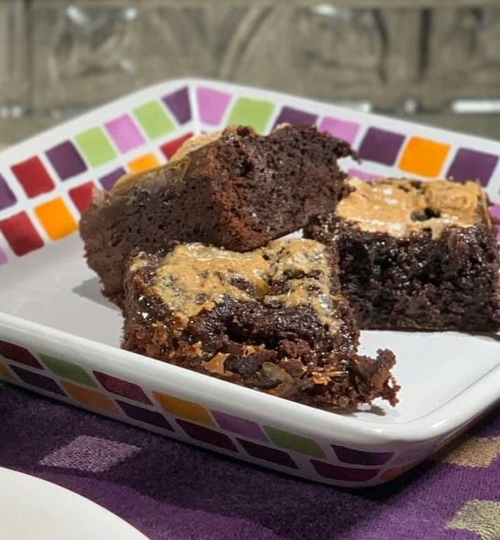 Marshmallow topped brownie on a white plate with more on a multicolored checked plate behind on a purple checked towel.