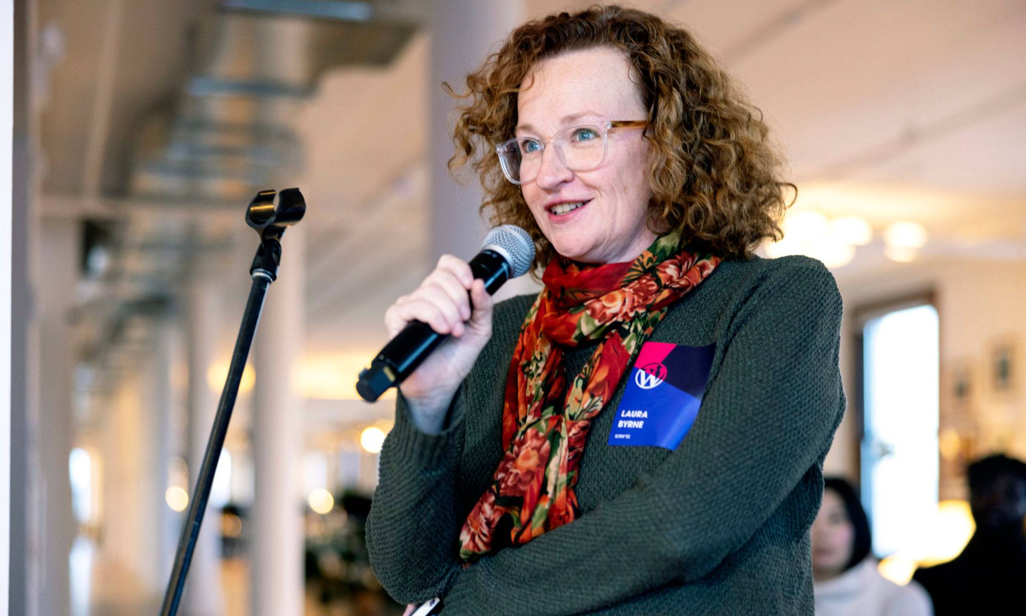 Laura Byrne, standing with a microphone in hand. The mic stand is in front of her. She's wearing a green sweater and colorful scarf. She has on glasses. Shee has brown/red currly hair