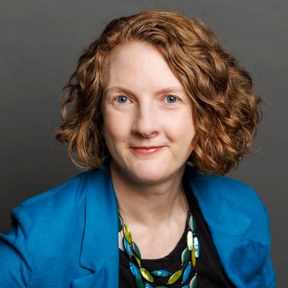 Kirsten is a white woman with shoulder length wavy red hair and blue eyes. She is wearing a black shirt, blue jacket, and blue and green necklace.