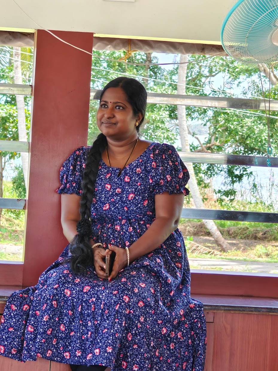 Jyolsna has long dark braided hair, a blue dress with pink flowers, and a lovely smile. She is holding a phone and wearing a necklace and bracelets. Behind her you can see trees through slatted windows.