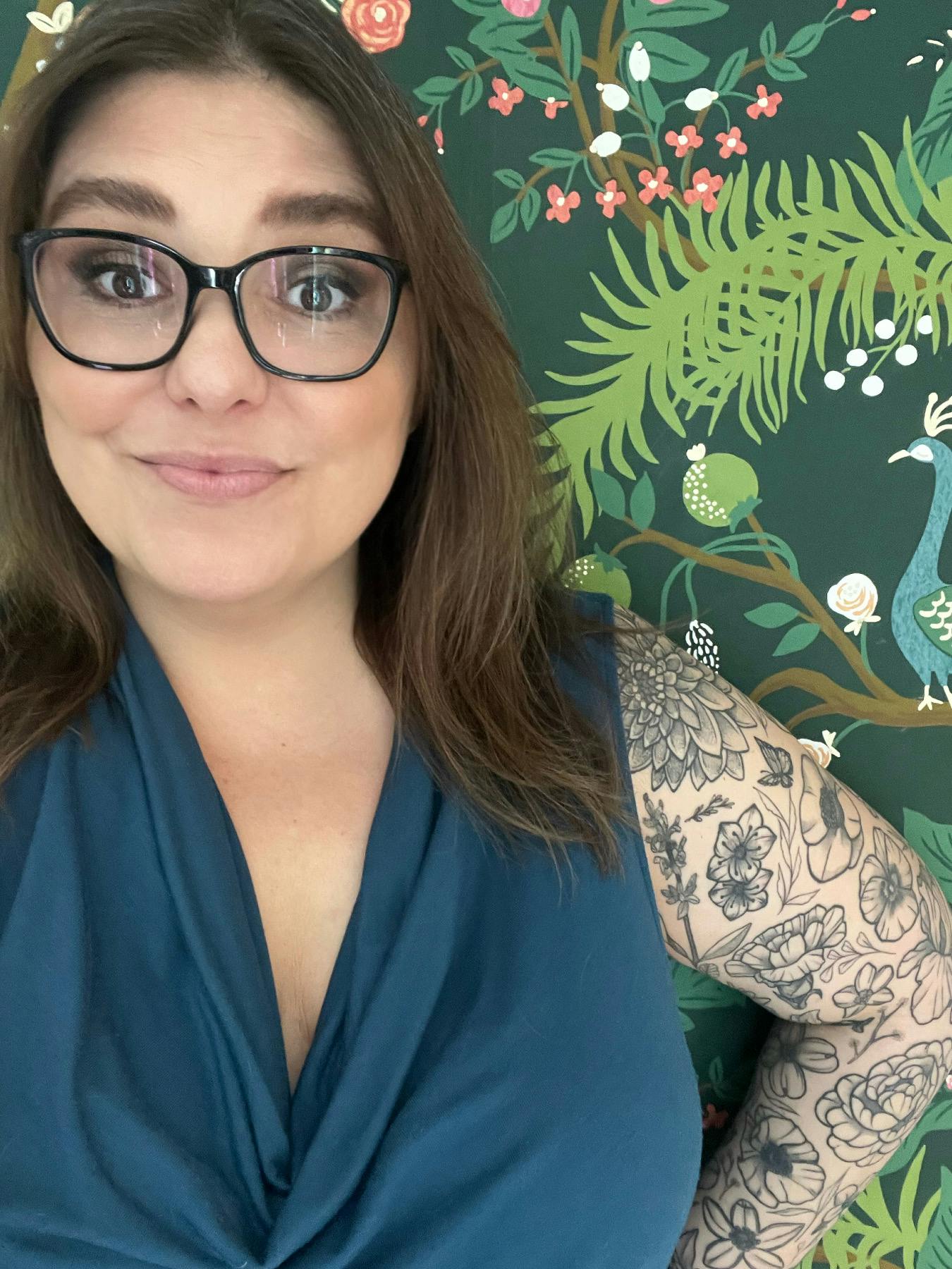 Jessica Frick, standing in front of a wall with floral motif. She has long brown hair, glasses, and a full sleeve of floral tattoos. She's wearing a teal blue shirt.