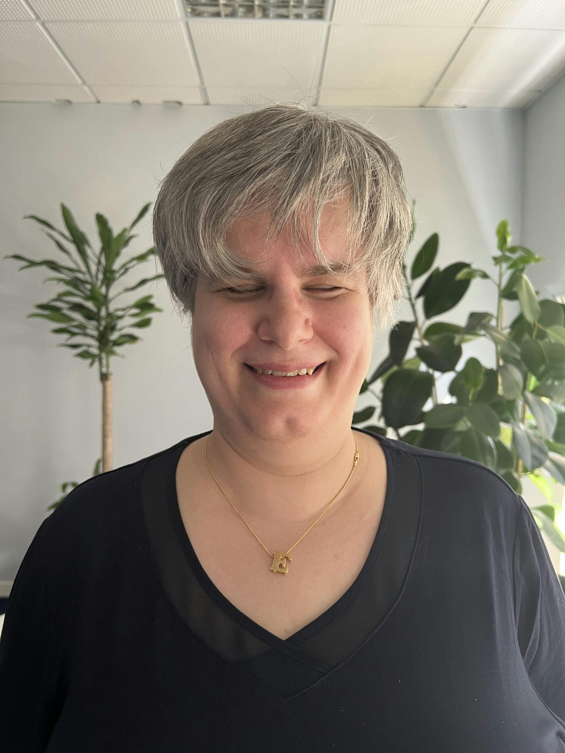 Elena Brescacin standing and smiling at the world. Wears a t-shirt and a golden necklace with a letter "E" pendant. She has short hair.