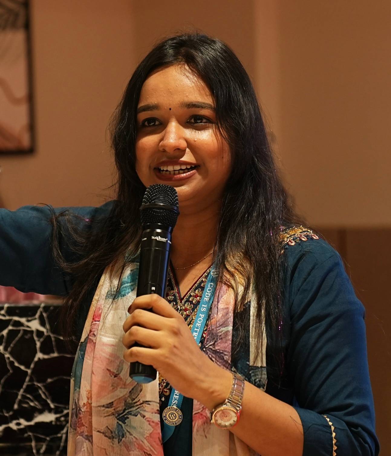 Astha Jain -  she has long dark hair with a warm smile. She's speaking into a microphone. 