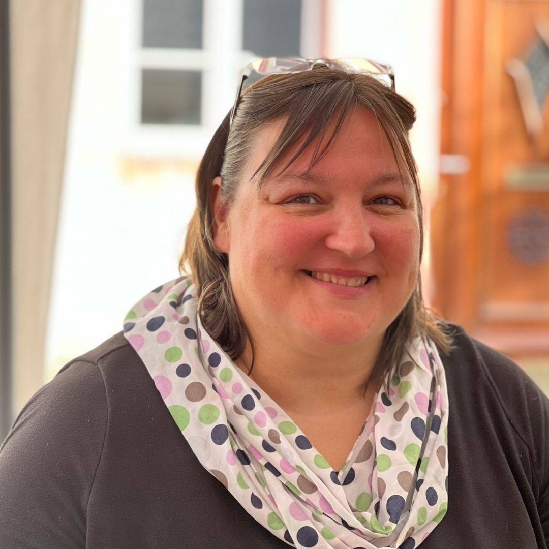 Birgit Olzen is a white woman with shoulder length light brown hair. She's smiling. She's wearing a grey shirt with a white scark with colorful polka dots. Her glasses are atop her head.