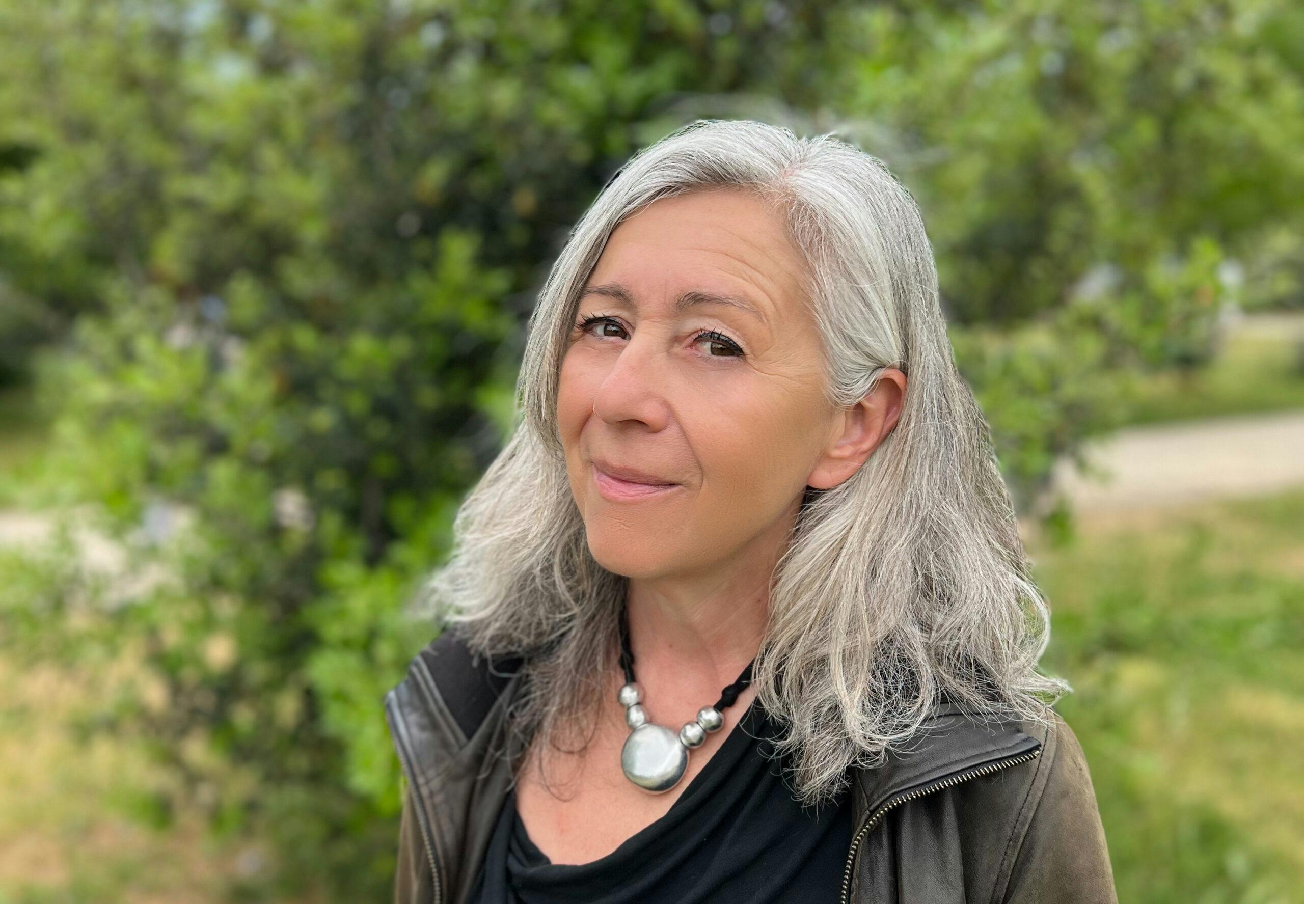 Piccia Neri is a woman with medium length greying hair, brown eyes, and a slight smile as she looks to the camera. She is wearing a black shirt, brown jacket, and silver necklace. There are trees in the background.