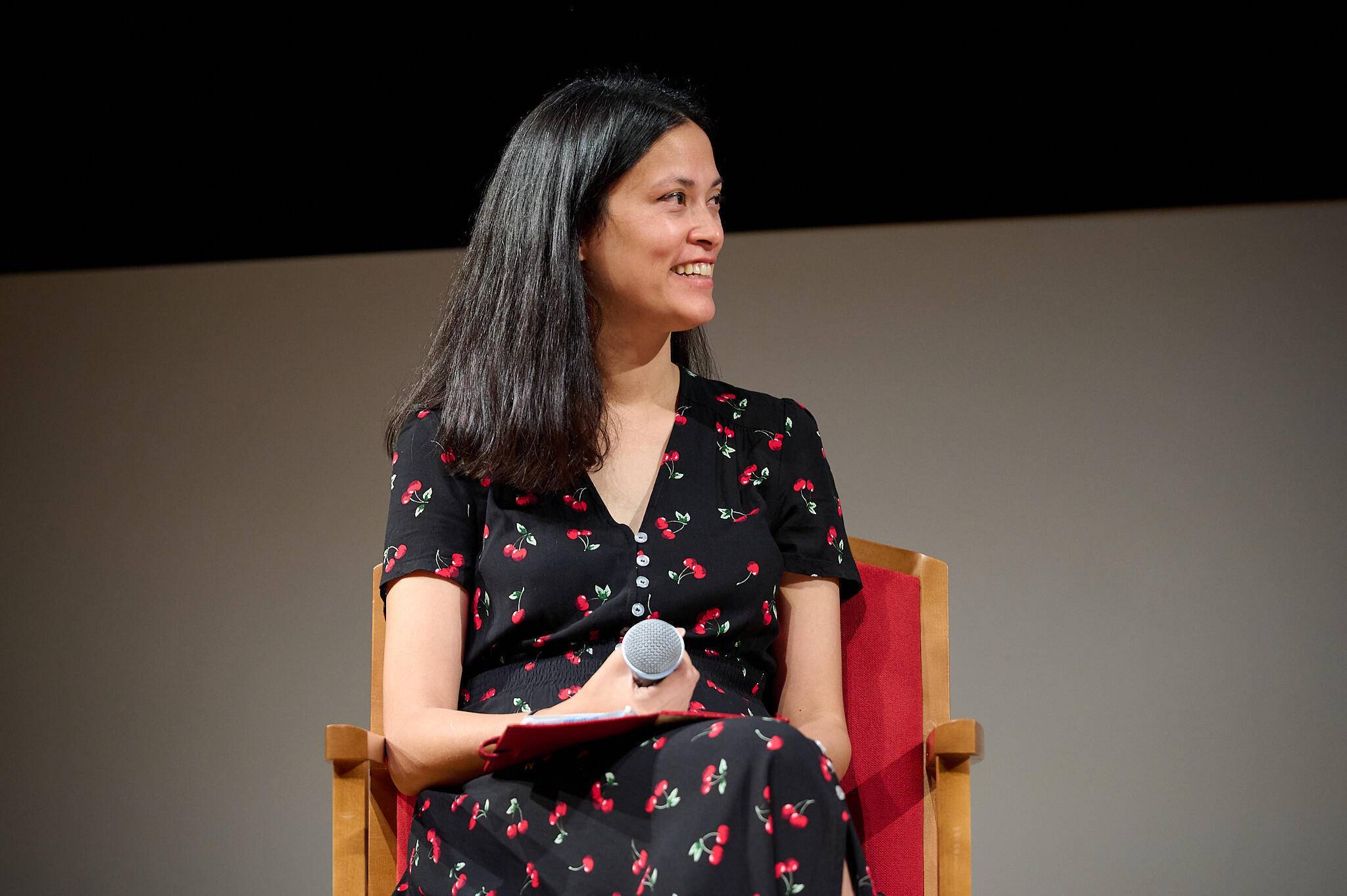 Josepha Haden Chomphosy sitting in a wooden chair with a microphone in her hand.