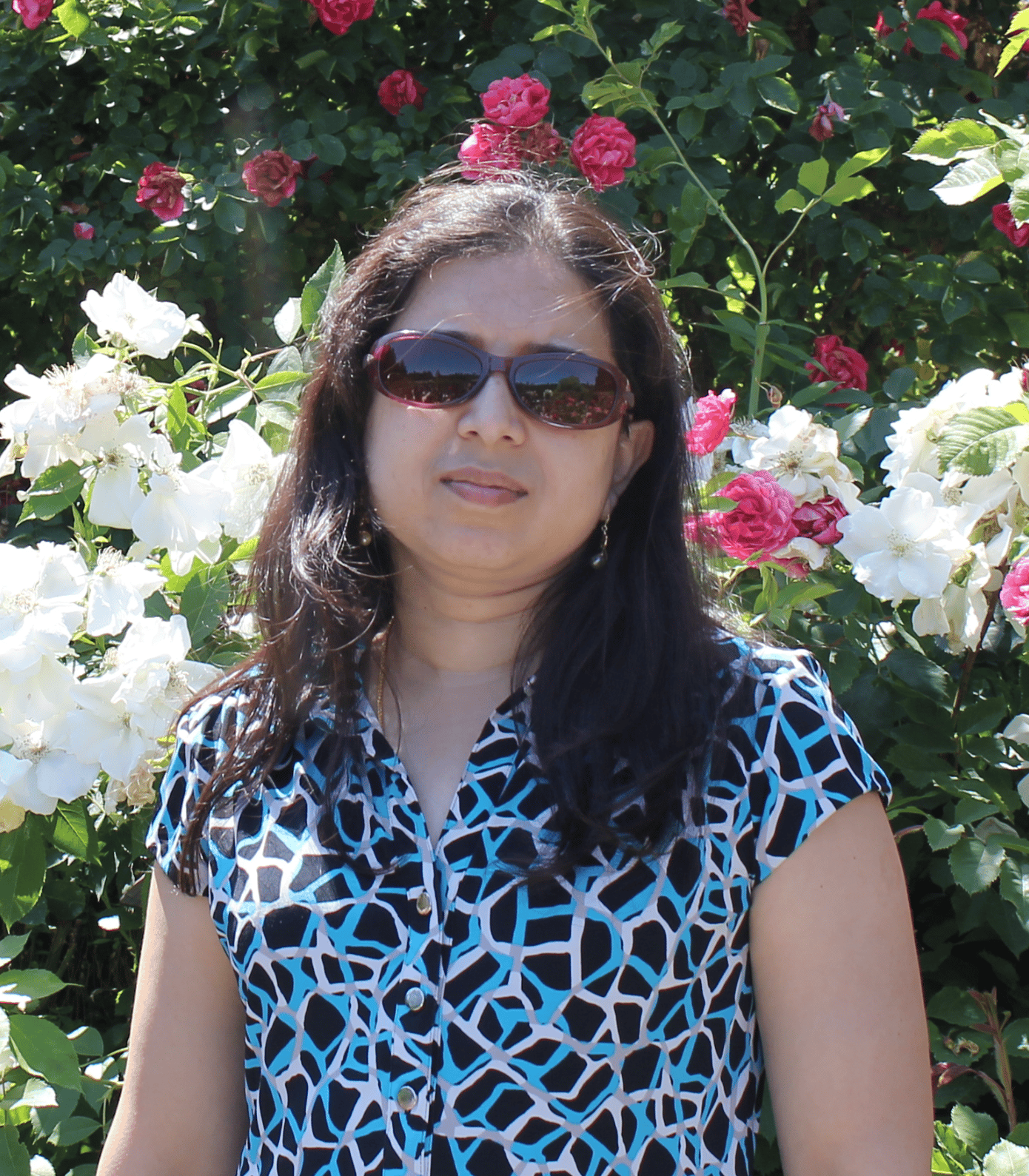 Chandrika Sista is an Indian woman with brown hair and sunglasses. She's wearing a black, blue, and white shirt and standing in front of a bush with green leaves and white and pink flowers.