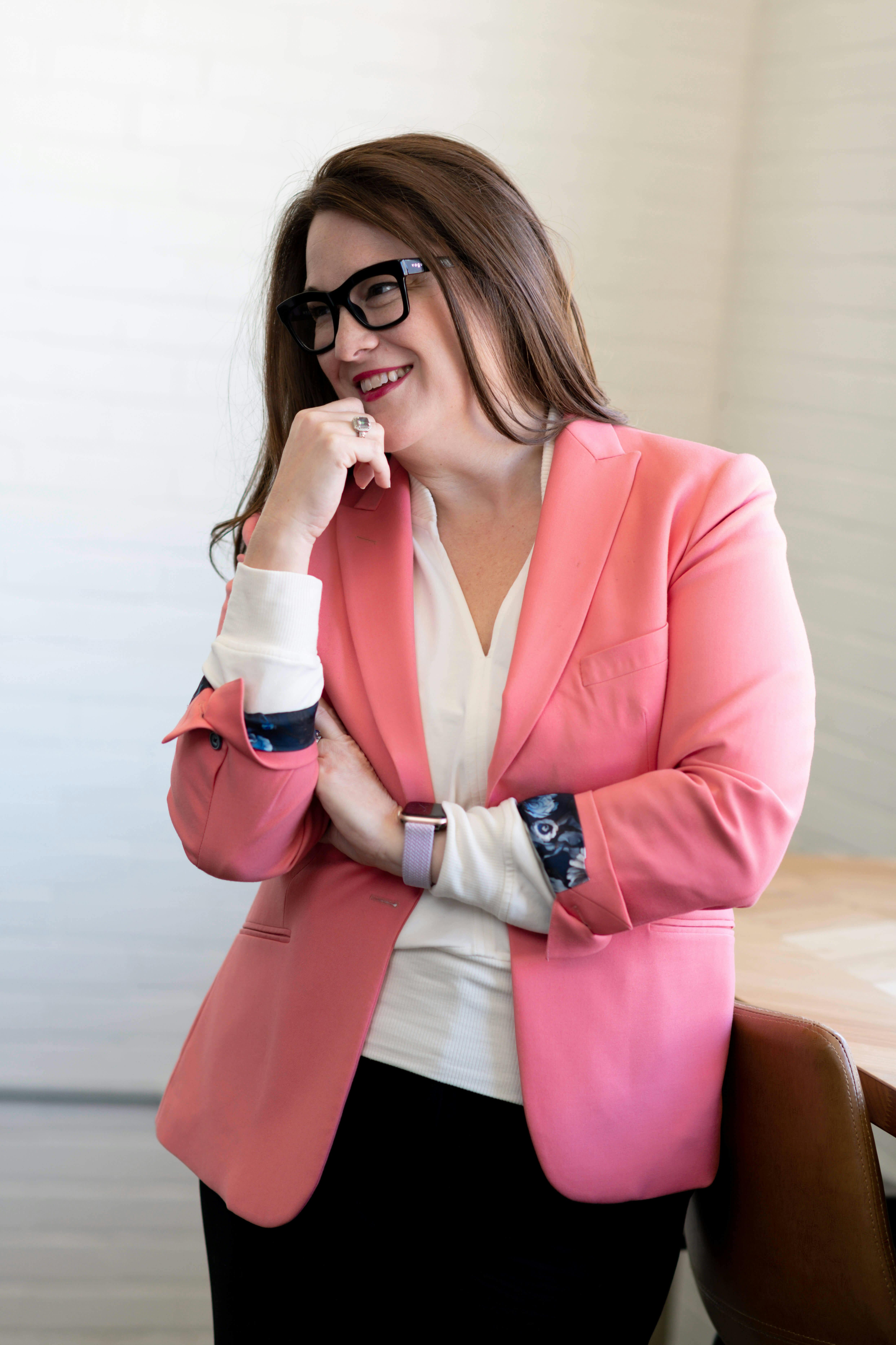 Lindsey Miller, in a coral colored blazer and white shirt, hair down, wearing black glasses and an Apple Watchh, smiling at something left of camera off screen