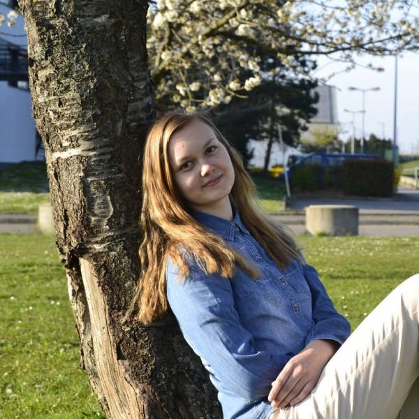 Lana has long light brown hair and a faint smile. She's sitting, leaning against a tree with her arms crossed. She's wearing a blue denim shirt and khaki pants.