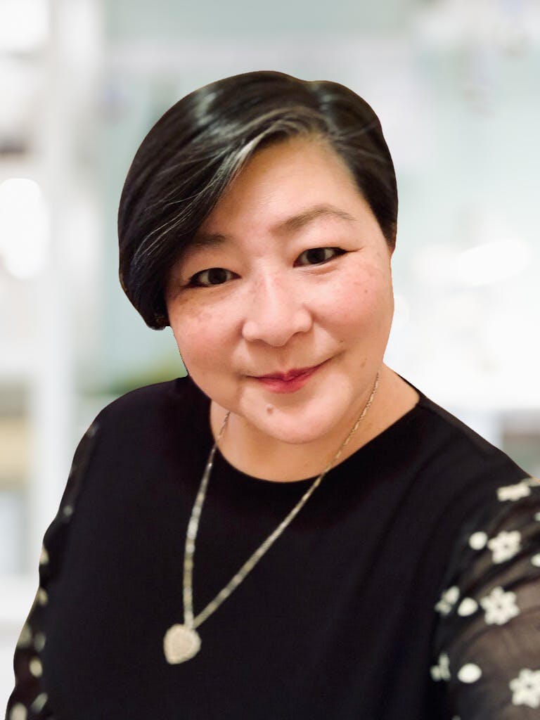 June Liu has short dark hair and is wearing a black shirt and hear-shaped necklace. She is against a blurred bright background.