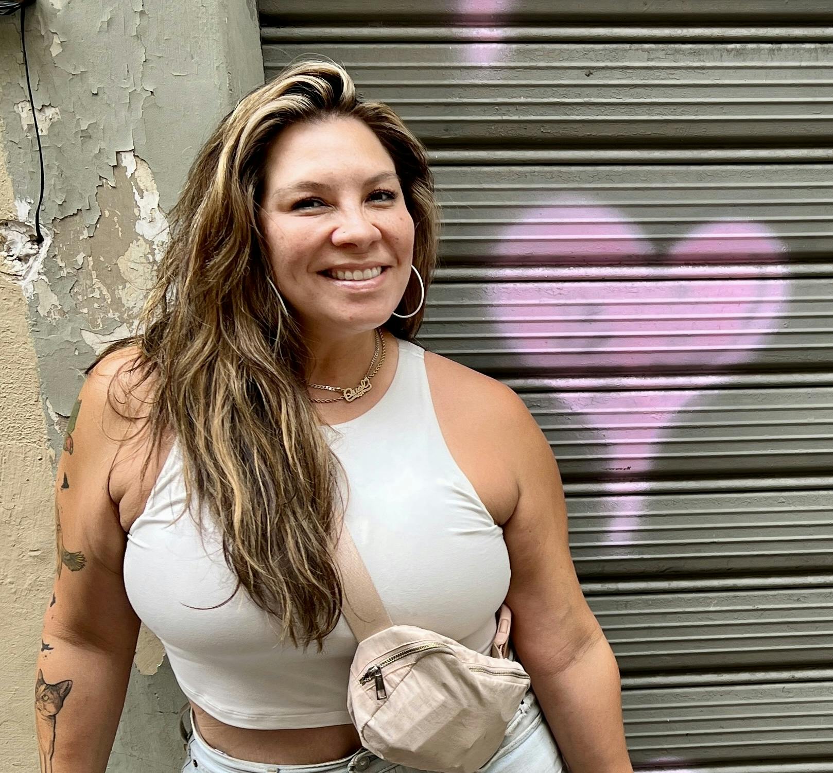 Raquel, standing in front of a wall with a pink heart painted on it. She has long brown hair, a big smile, hoop earrings, and is wearing a white top and beige bag..