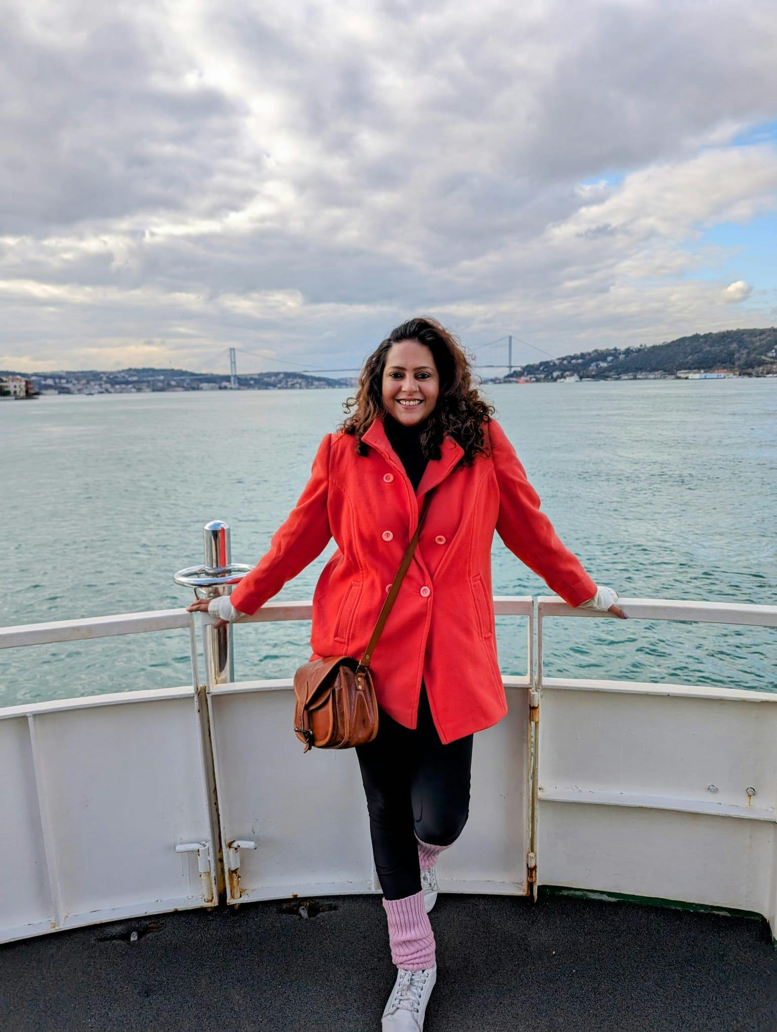 Chandni Ssirohi is an Indian woman with curly brown hair and a big smile. She is wearing black pants, a bright red coat, and a brown bag. She's standing on a boat with her back to the water. There is a bridge in the background.