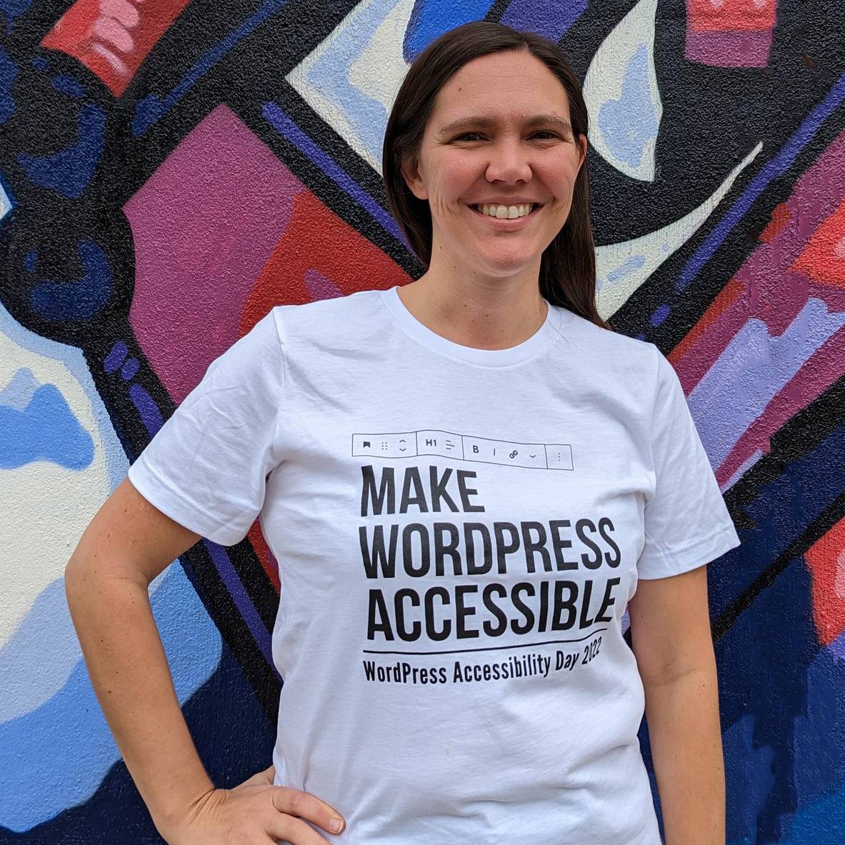 Amber Hinds standing in front of a colorful abstract wall, wearing a white t-shirt that says "make wordpress accessible: WordPress Accessibility Day 2022. She has long brown hair and a big smile. She's standing with one hand on her hip.