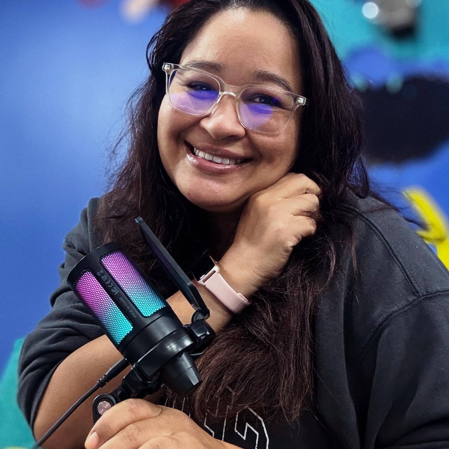 Lua is a smiling woman with long dark hair and glasses. She is wearing a dark sweatshirt and holding a microphone.