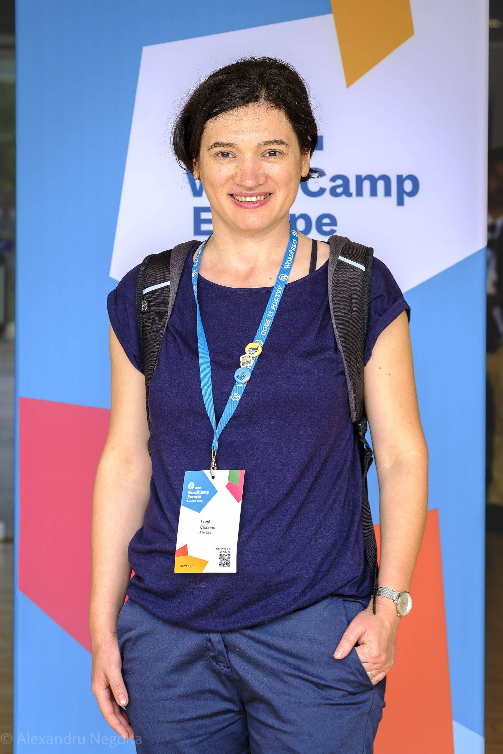 Lumi Ciobanu is a white woman with dark hair tucked behind her ears. She is wearing a dark blue shirt, WordCamp Europe badge, and a dark backpack. She has brown eyes and a lovely smile.