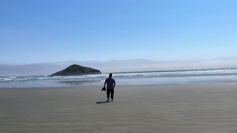 A person walking along a sandy beach towards a large rockk.