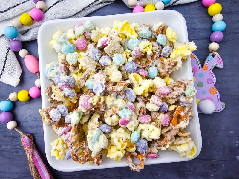 Overhead view of a square bowl of Bunny Bait Snack mix with popcorn, pretzels, white chocolate and pastel colored M&Ms