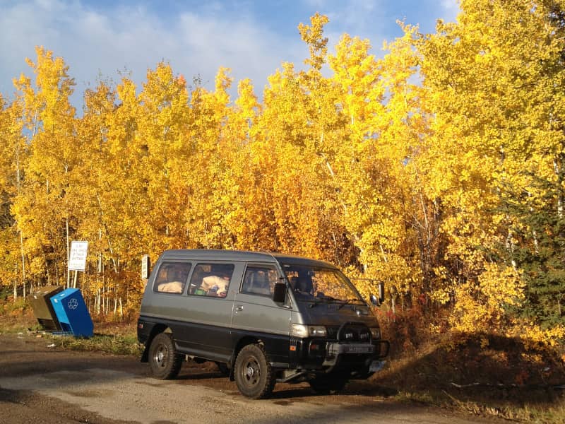 a Delica van at a rest stop with the leaves of Fall trees behind it a bright yellow. 