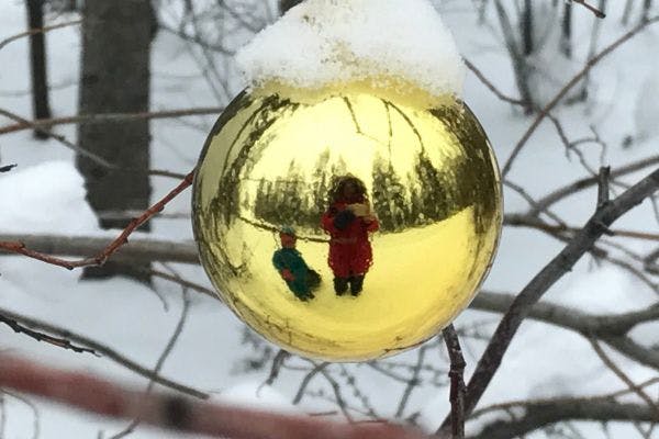 Mom & daughter in winter suits reflected in an ornament hanging on snowy branches