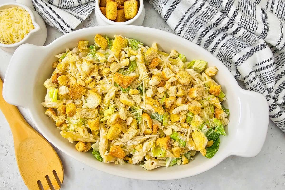 An overhead view of an oval bowl of Caesar Pasta Salad, with wooden serving spoon beside it. 