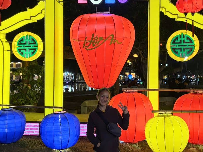 A young girl smiling and giving the peace sign in front of a display of lit up lanterns at night, with the words Hoi An on them