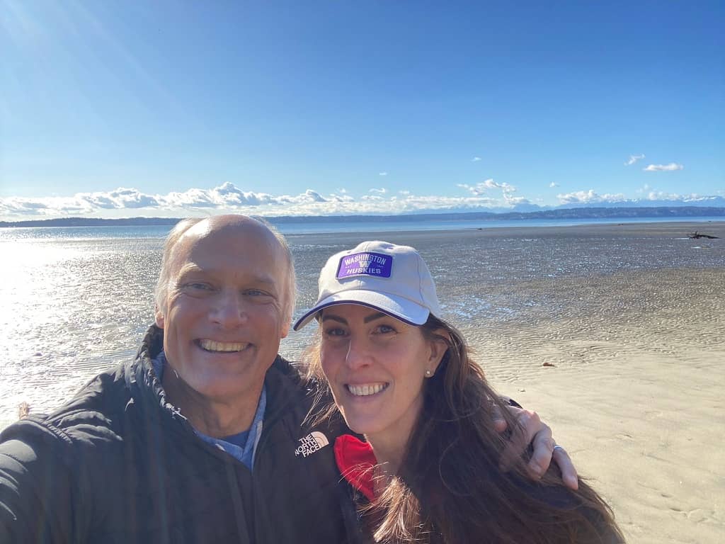 A smiling couple takes a selfie on a wide, sandy beach at low tide. The ocean stretches out behind them under a bright blue sky with scattered clouds. One person wears a dark jacket, the other a light-colored baseball cap, and both appear relaxed and happy in the sunny coastal setting.
