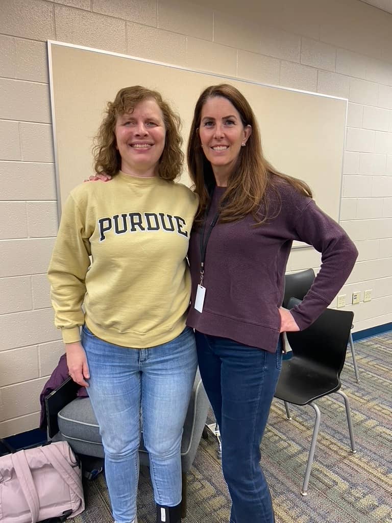 Two women stand side by side in a classroom or meeting room, smiling at the camera. The woman on the left has curly light-brown hair and is wearing a light yellow Purdue sweatshirt and blue jeans. The woman on the right has long brown hair and is wearing a dark plum long-sleeve top and blue jeans, with one hand on her hip. Behind them are stackable black chairs, a whiteboard on a cinderblock wall, and a light-colored jacket and bag resting on a chair, giving the impression of an informal talk, workshop, or school setting.