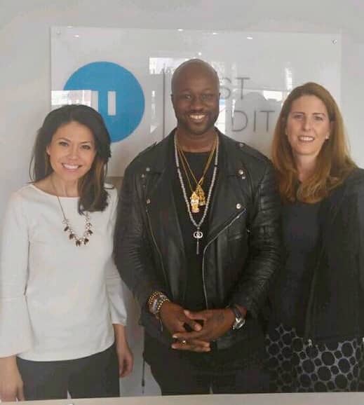 Three people stand in front of an acrylic sign with a blue pause button and writing just behind their heads. - The woman on the left is attractive with a lovely smile, long shoulder-length hair, a white top, and black pants with a chunky beaded necklace. - The African American man is in the middle wearing a black leather moto jacket with several chains around his neck. - The third person is a Caucasian female with light brown shoulder-length hair. She is smiling in a dark top and a gray patterned skirt.