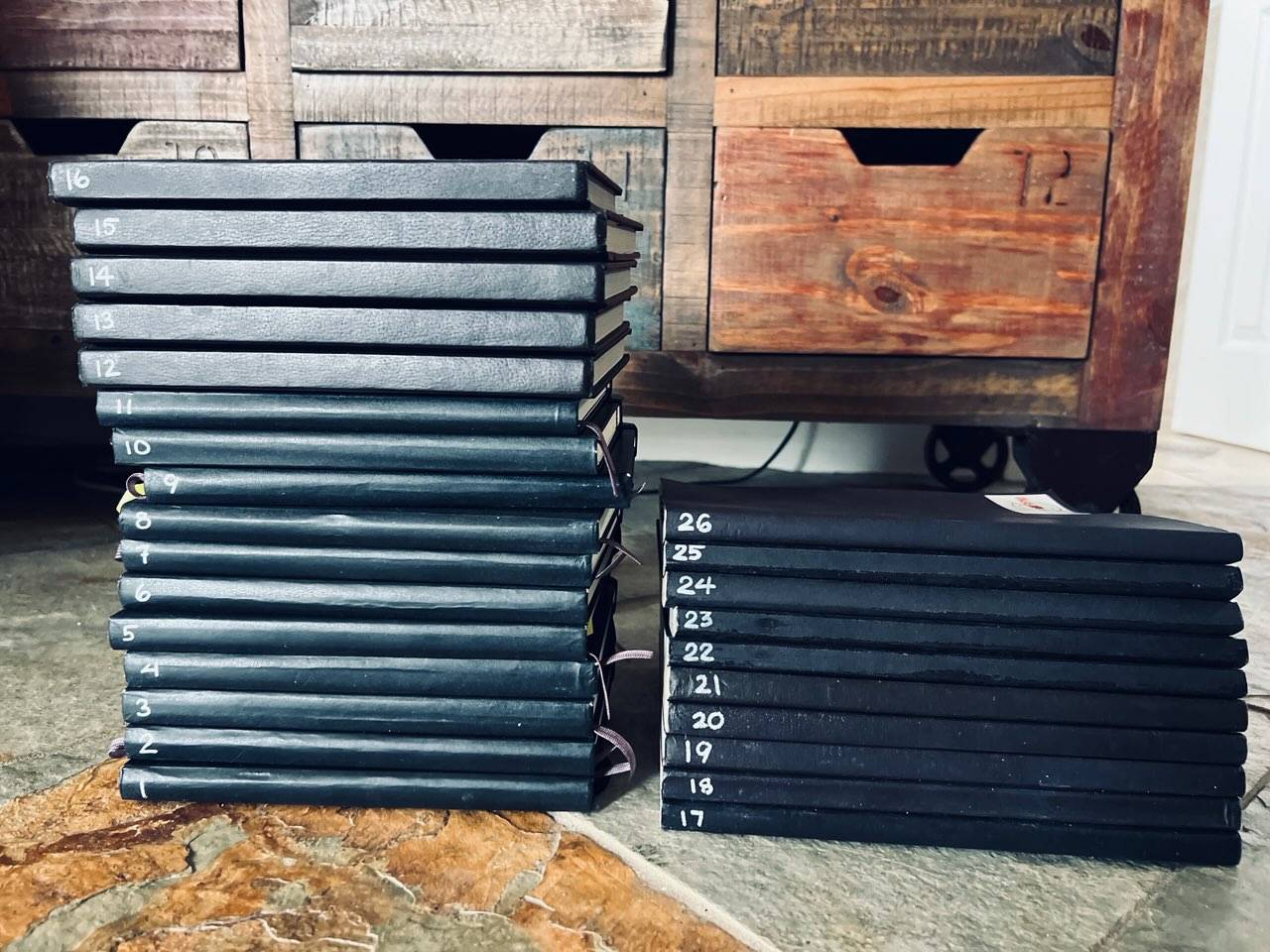 Two stacks of black notebooks sit side by side on the floor in front of a wood chest. The floor is stone gray and terra cotta. The books are numbered. The left stack of books starts at the bottom (book 1) going to 16. The right stack of books starts at 17 on the bottom going to 26. 
