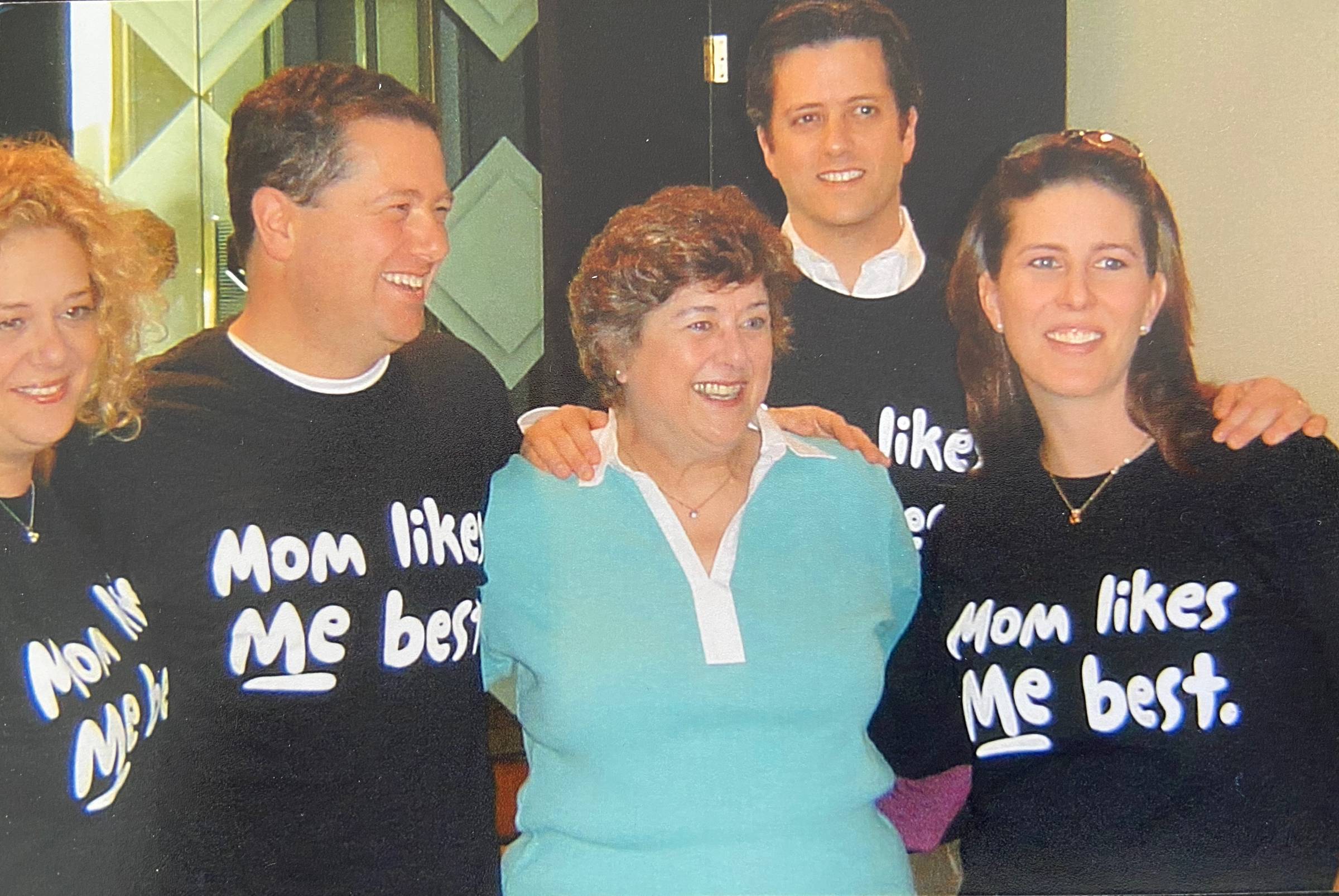 A family group stands closely together indoors, smiling and laughing. A woman in a light blue sweater stands at the center, surrounded by four adult children with their arms around her shoulders. Several of them wear black shirts with the playful phrase “Mom likes me best,” capturing a warm, affectionate, and humorous family moment.