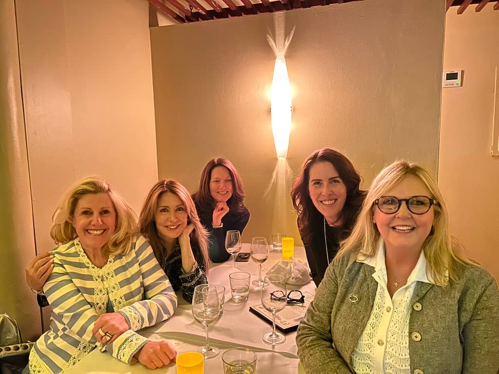 Five women seated around a restaurant table smiling at the camera during a dinner gathering. Two women sit in the front, two sit behind them, and one sits at the center back. The table has wine glasses, water glasses, notebooks, and small yellow candle-like lights. The group appears relaxed and happy, with warm lighting and a modern wall light fixture glowing behind them.