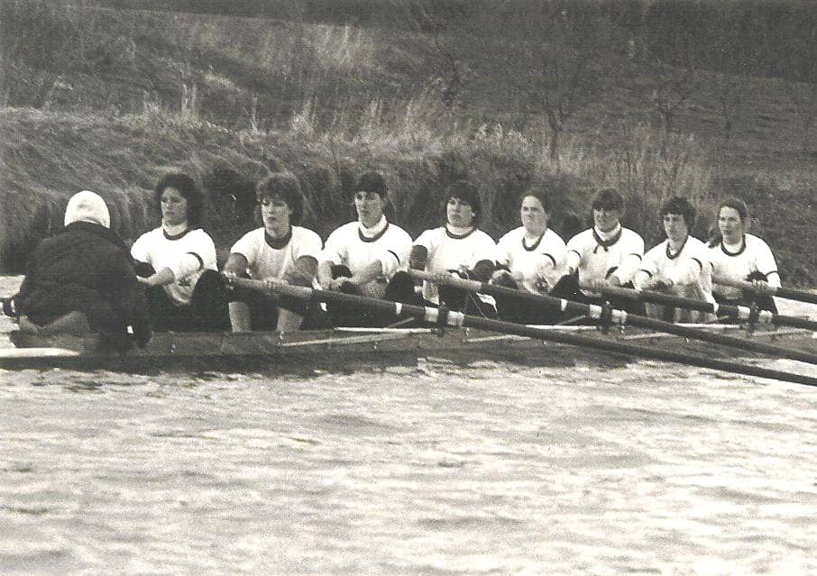 A black and white photo of the women's Varsity 8 sitting at start in the catch position ready to row. They are in race white t-shirts. The coxswain's back is to the photographer wearing a black jacket and a white hoodie sweatshirt on her head. The water is rough, you can see the riverbank on the left side of the photo. I sit in the sixth seat. 