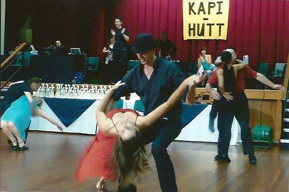 Amy in a red strappy dress in a dip, on her back, head towards the camera, as Chris dressed all in black with a black hat holds her with outstretched arms. Amy has her right leg bent as support and her left leg extended. Her right arm is up over her head and her left is down resting on Chris' right arm which is holding her left hip.