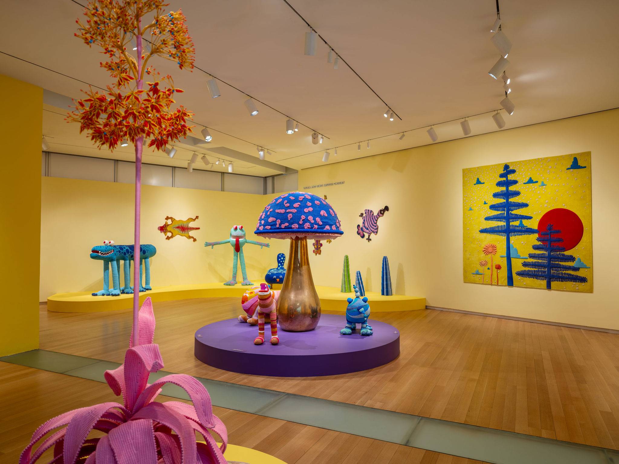 A wide-angle installation shot of the vibrant Haas Brothers "Uncanny Valley" museum exhibition, featuring whimsical, brightly colored creature sculptures and a large patterned mushroom on a purple platform against bold yellow walls.