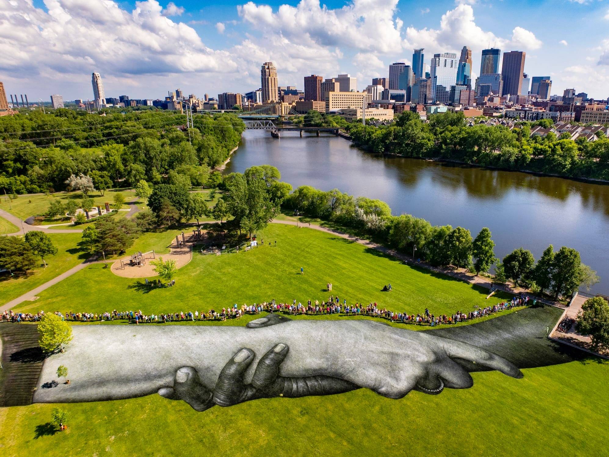 High-angle aerial view of a massive black-and-white land art mural by Saype at Boom Island Park, depicting two intertwined hands painted on the grass. The Mississippi River and downtown Minneapolis skyline form the background under a partly cloudy sky.