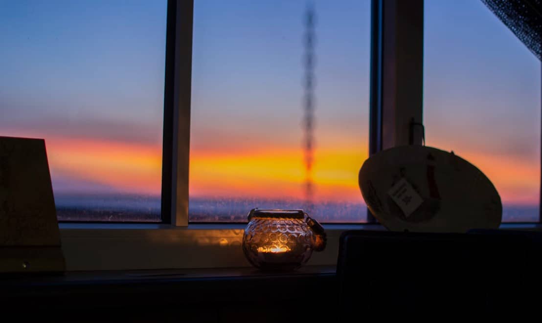 Image of a candle in a glass, with a window showing v blurry sunset over water