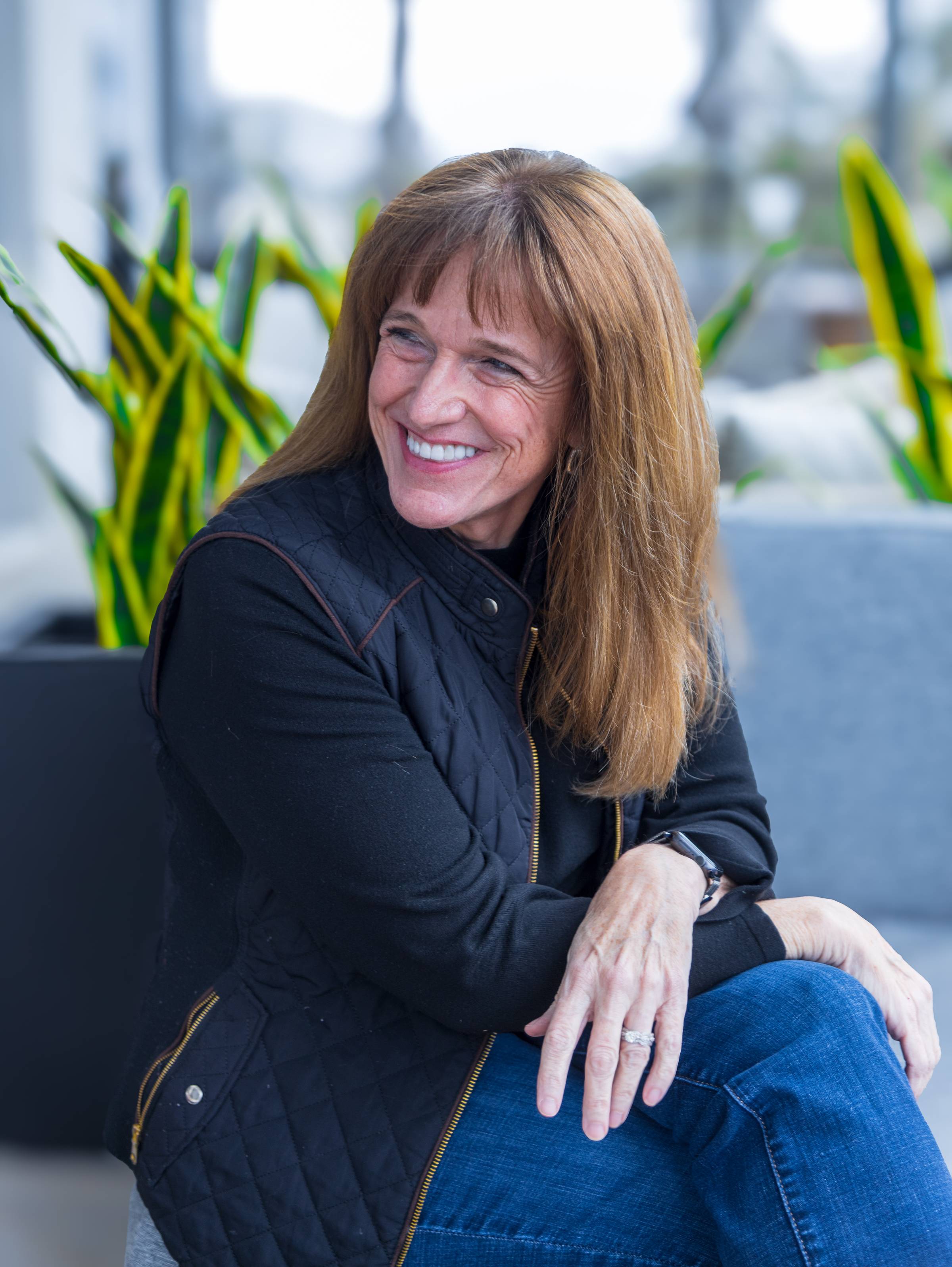 A woman with long brown hair smiles while seated outdoors. She is wearing a black quilted jacket and blue jeans, with her hands resting on her knee. In the background, there are green leafy plants. The atmosphere appears casual and relaxed.